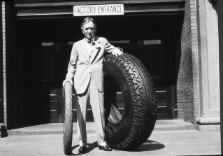 Harvey Firestone Sr. standing at the Akron, Ohio, factory entrance with an original Firestone non-skid tread tire and a large tire, ca. 1926-1930.